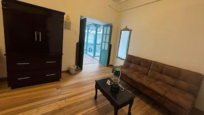 Une pièce avec un plancher en bois, un canapé marron, une petite table sur laquelle se trouve une bouteille verte, une armoire en bois foncé et une porte qui donne sur l’extérieur.