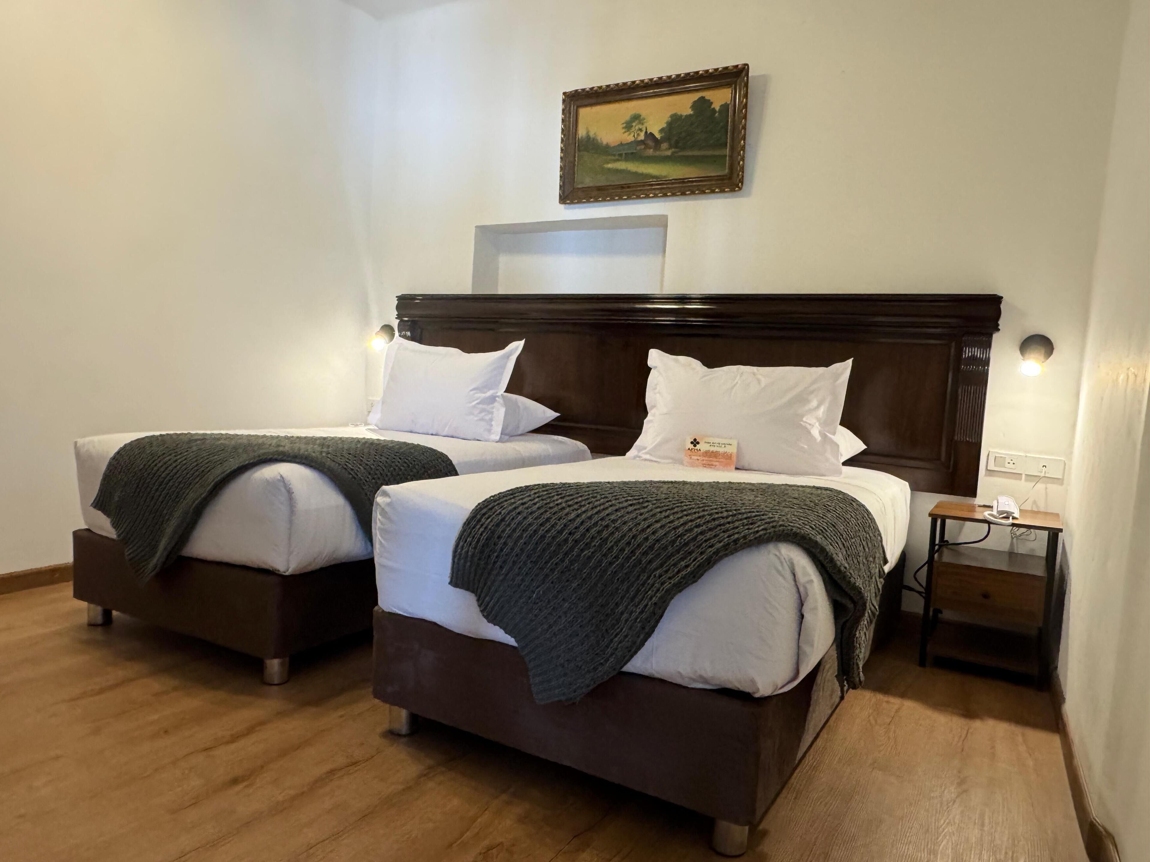 Een hotelkamer met twee bedden, elk met witte lakens en een donker hoofdeinde. Daarboven hangt een lijst met een landschapsbeeld, een nachtkastje met een lamp en een klein nachtkastje met een telefoon.