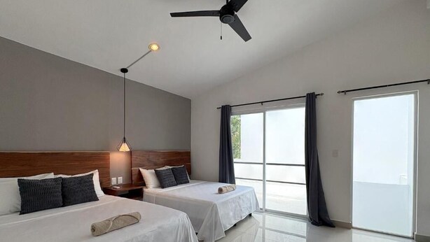Une chambre d’hôtel avec deux lits, un ventilateur de plafond, une grande fenêtre avec des rideaux et une table de chevet avec une lampe.