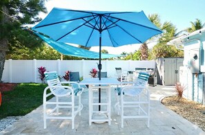 Een terras buiten met een tafel en stoelen, een blauwe parasol en een witte hek in de achtergrond.