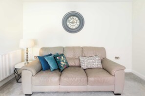En beige lädersoffa med tre kuddar, en rund väggklocka, ett litet bord med en lampa och ett element.
