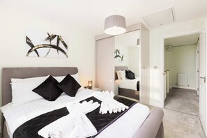 En snyggt bäddad säng med vita och svarta sängkläder, en grå sänggavel och en svart, vadderad pläd. En vit handduk är prydligt vikt och ligger på sängen, och en modern hänglampa hänger ovanför.