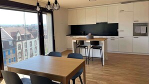 A modern kitchen with white cabinets, a wooden dining table, and black chairs. There is a large window providing a view of buildings outside. - Top modern Apartment near Novartis Campus (Basel)