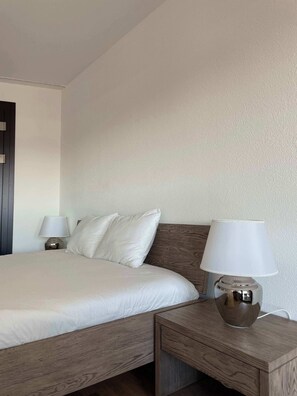 Ein ordentlich bezogenes Bett mit weißen Leinen, einem Holzkopfstück und einer Nachttischlampe mit weißem Lampenschirm.