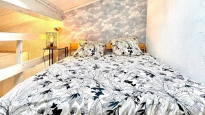Une chambre avec un plafond en pente, un lit recouvert d’une couvre-lit à motifs floraux, une fresque murale représentant des nuages et une petite table avec une lampe.