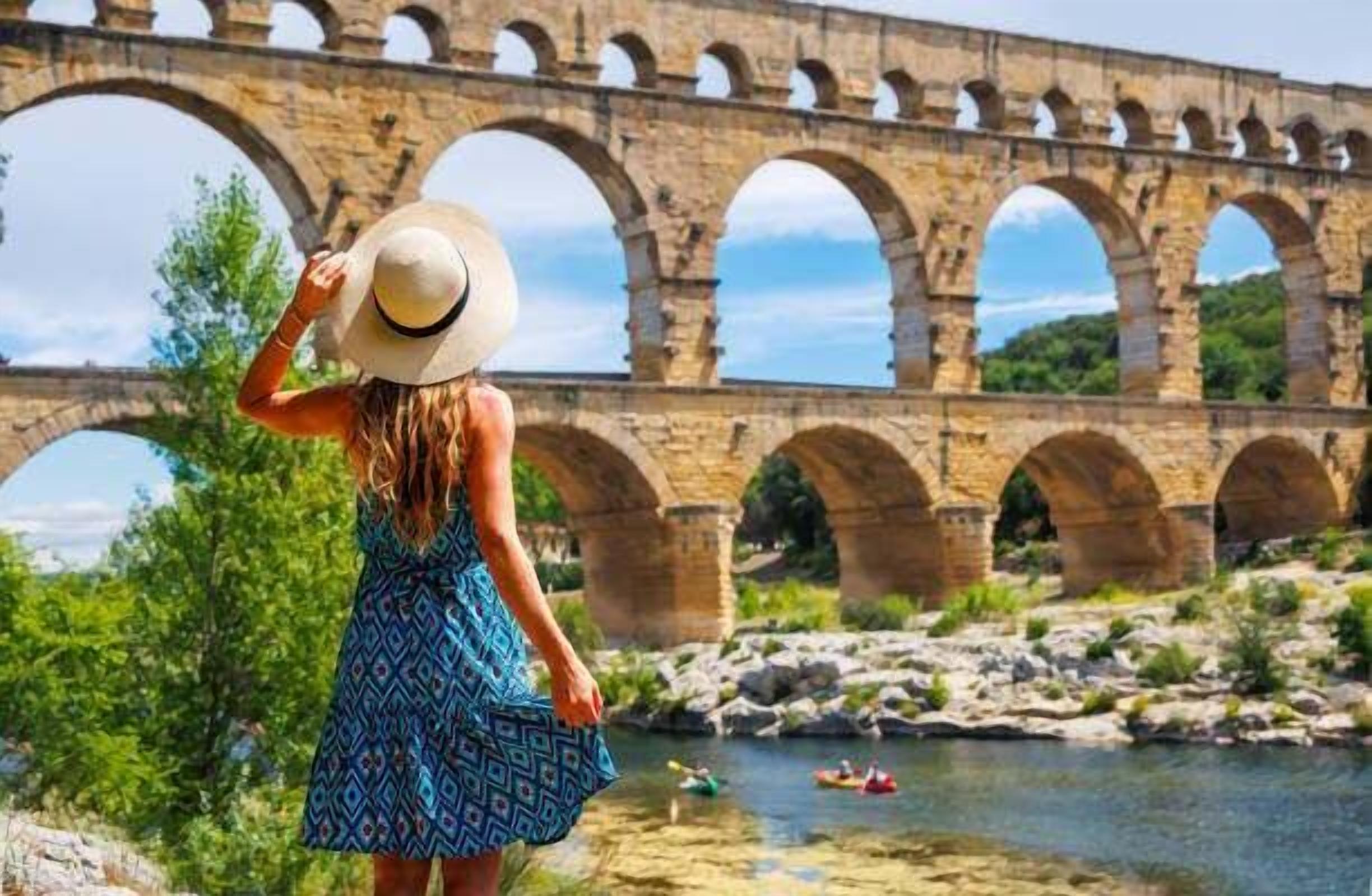 Une personne, vêtue d’une robe et d’un chapeau à motifs, se tient face à un pont de pierre à plusieurs arches qui enjambe une rivière. On aperçoit, en arrière-plan, de la verdure et un ciel dégagé.