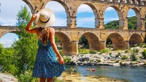 Une personne, vêtue d’une robe et d’un chapeau à motifs, se tient face à un pont de pierre à plusieurs arches qui enjambe une rivière. On aperçoit, en arrière-plan, de la verdure et un ciel dégagé.