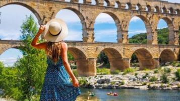 A person in a patterned dress and hat overlooking a stone bridge with multiple arches over a river, with greenery and a clear sky in the background.