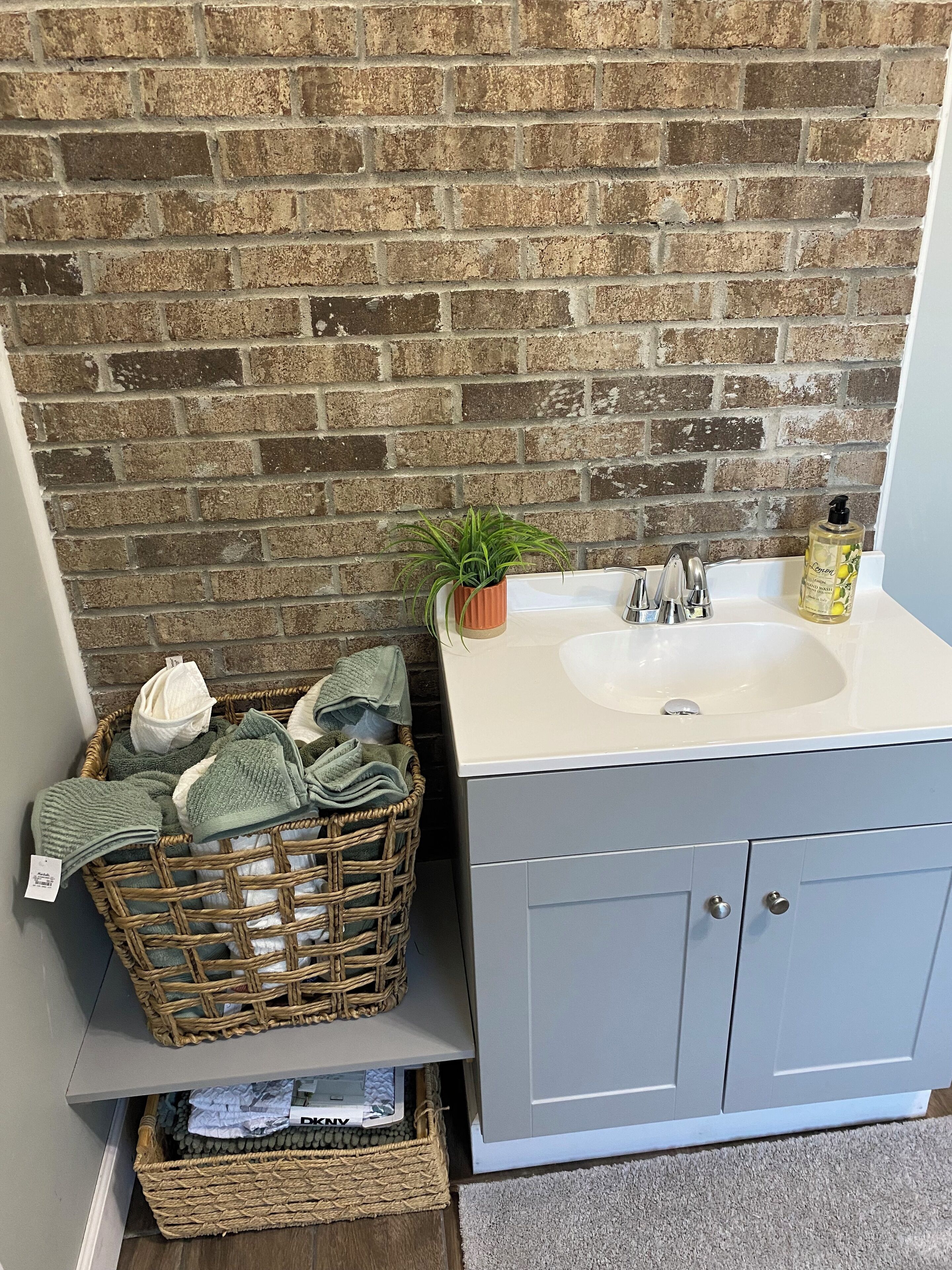 Une salle de bain avec un lavabo blanc, un mur en briques, un panier en osier contenant des serviettes et une petite plante en pot.