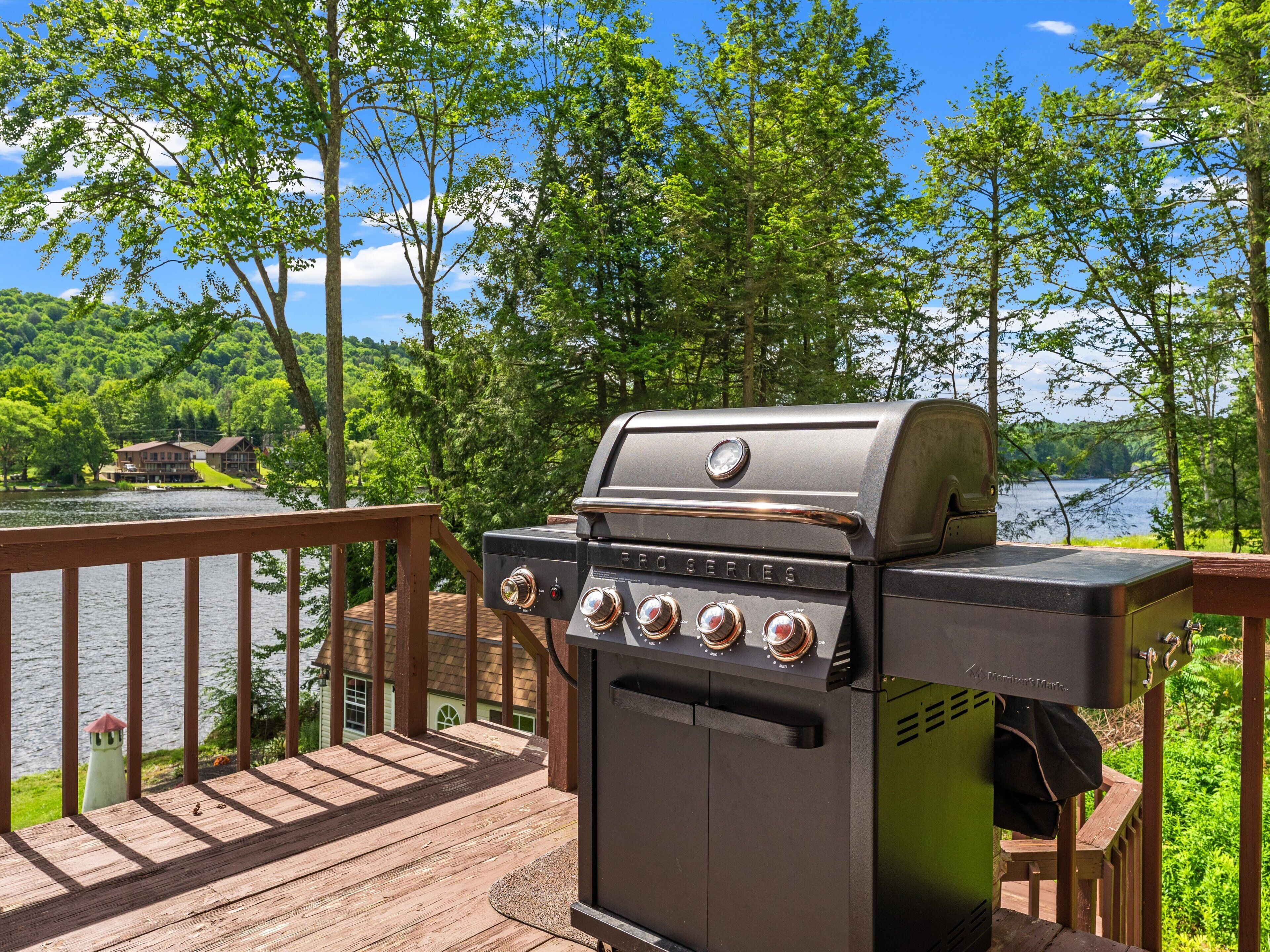 A grill on a wooden deck overlooking a lake with trees and a house in the background.