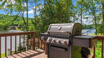A grill on a wooden deck overlooking a lake with trees and a house in the background.