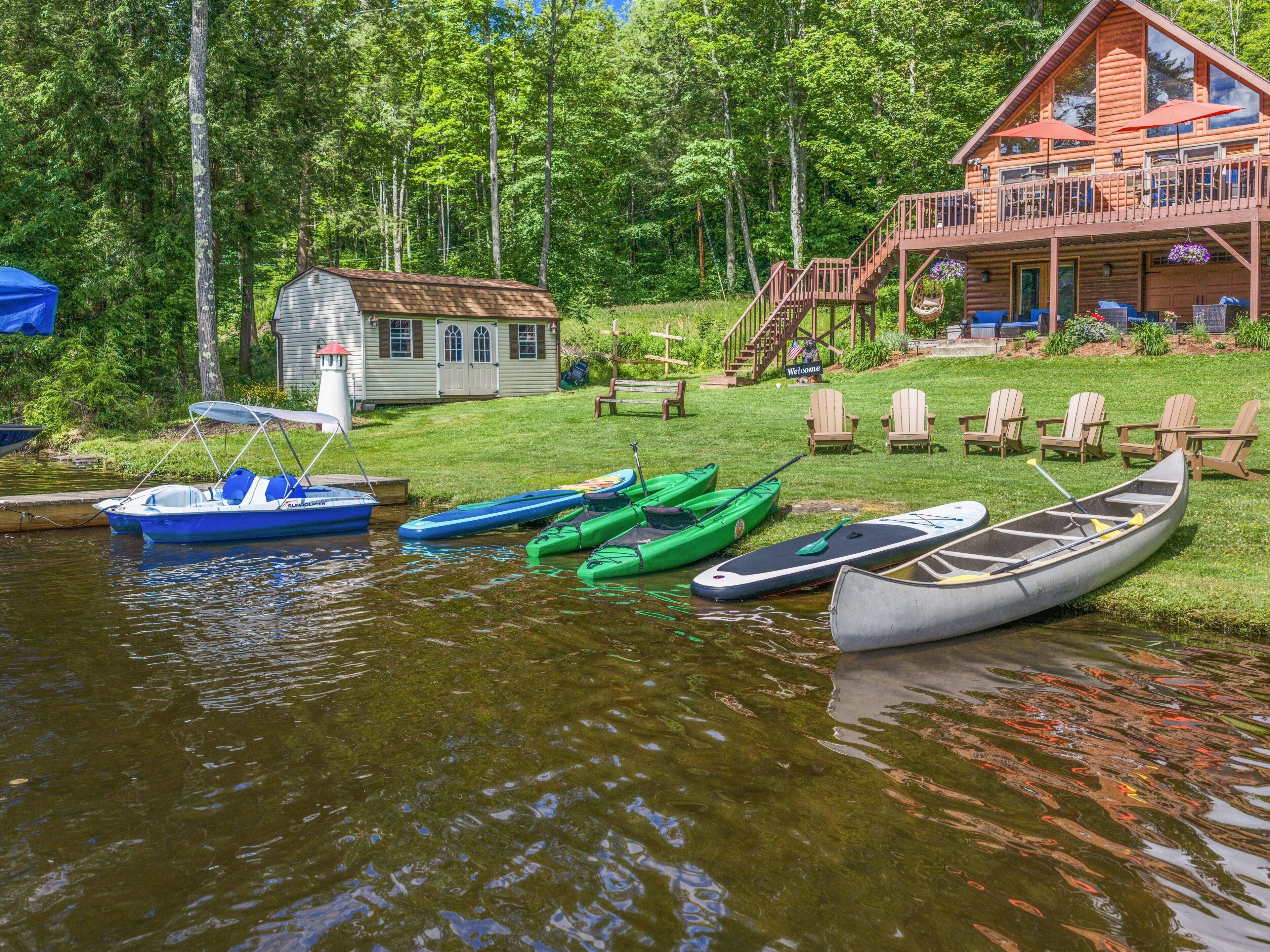 A lakeside cabin with a deck, boats, and a small house in the background.