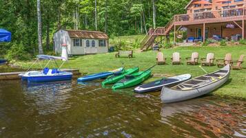 A lakeside cabin with a deck, boats, and a small house in the background.