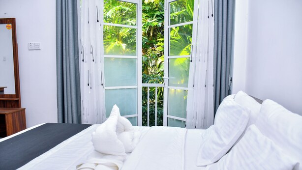 A neatly made bed with white linens, a wooden nightstand, and a large window with curtains. - Eco Bless Inn (Kandy)