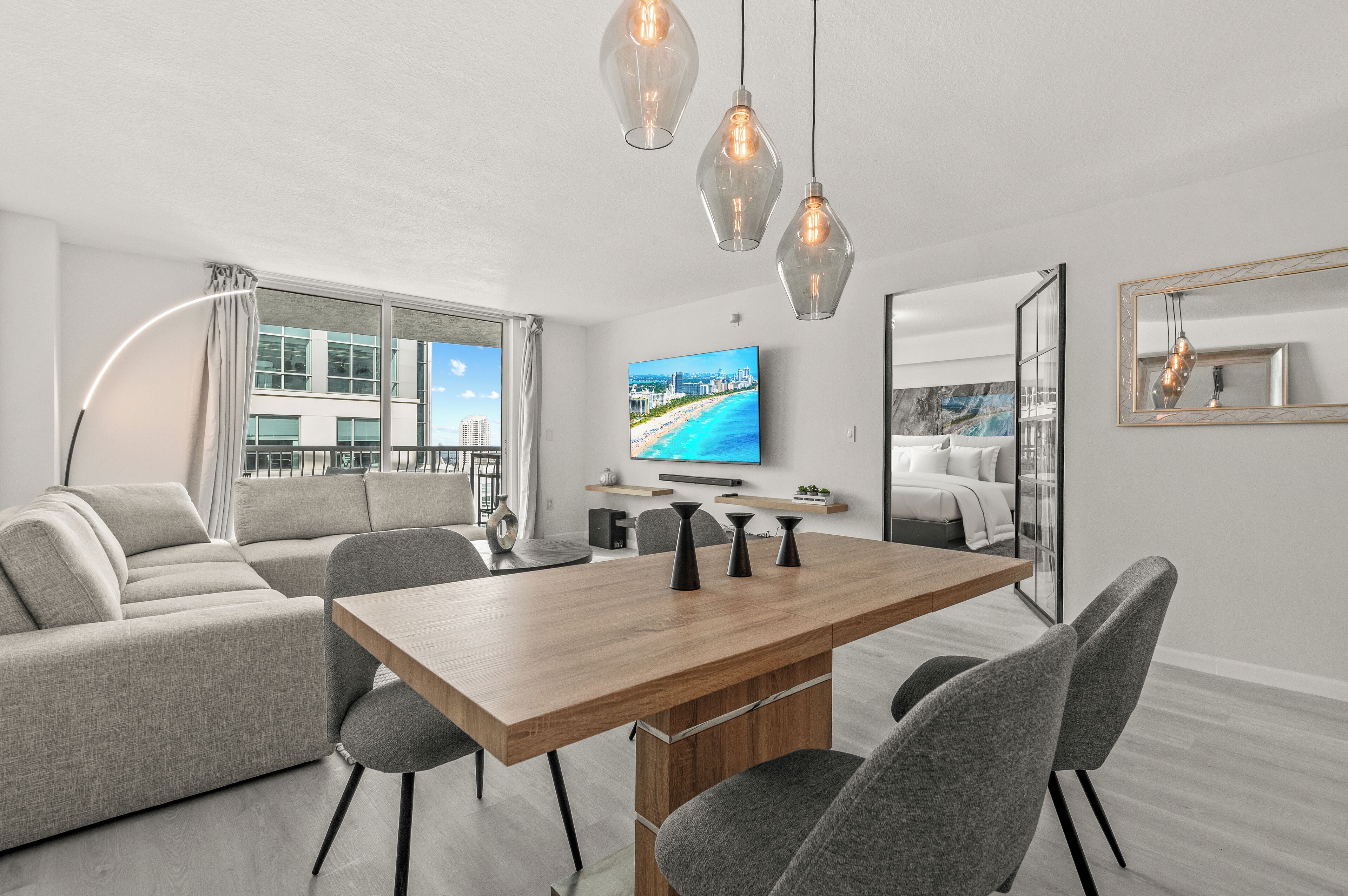 Een moderne woonkamer met een houten eettafel, grijze banken en een televisie aan de muur.