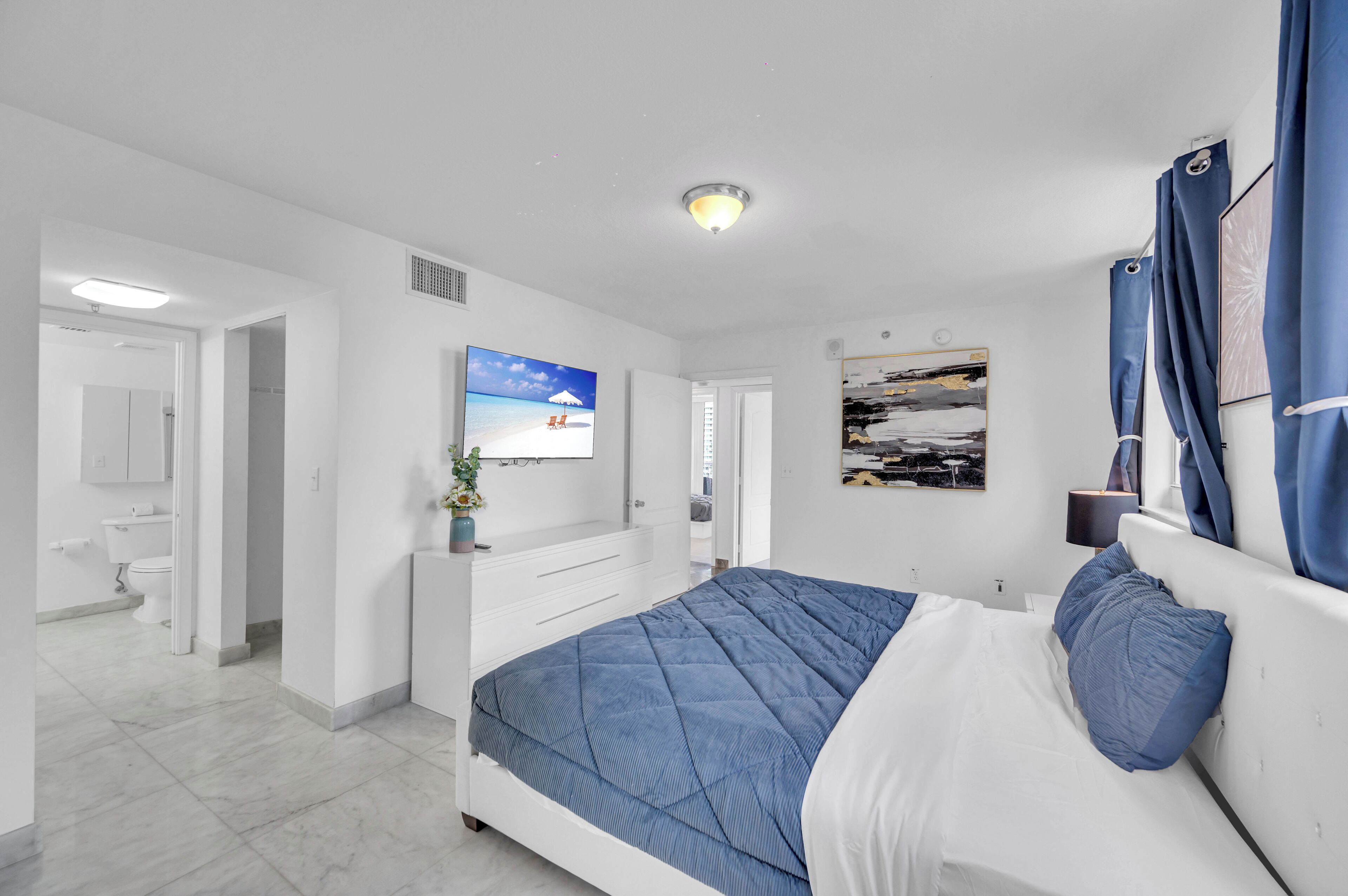 Ein modernes Schlafzimmer mit Bett, Fernseher, einem Gemälde und einem sichtbaren Badezimmer im Hintergrund.
