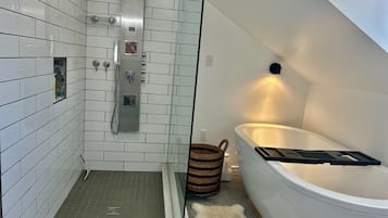 Un baño moderno con paredes de azulejos blancos, ducha con panel de control, bañera con luz y una cesta tejida.