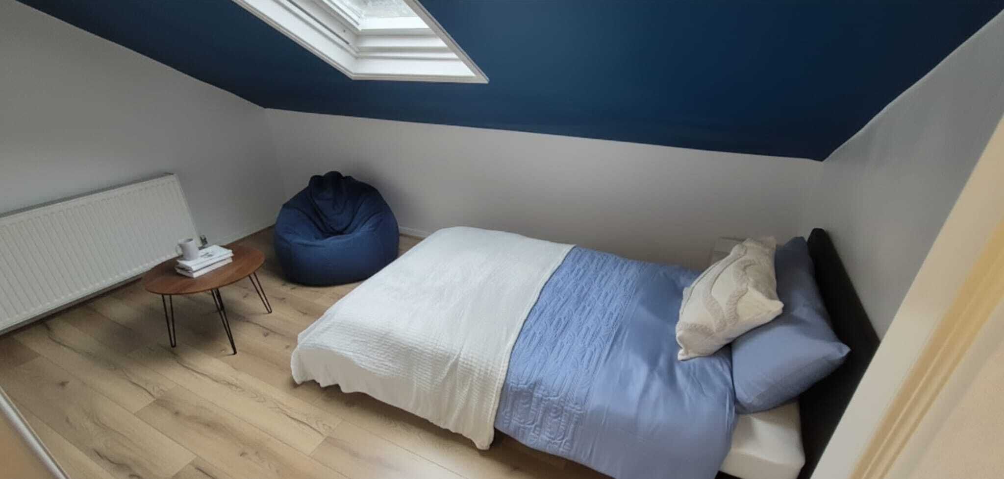 Un dormitorio con cama, sillón de plumas, una mesita y un tragaluz.