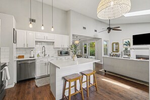 Une cuisine moderne avec des meubles blancs, un îlot central, des tabourets de bar, un ventilateur de plafond et un téléviseur.