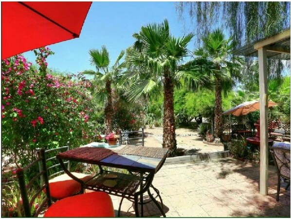 Ein Patio im Freien mit Tischen und Stühlen, umgeben von Palmen und blühenden Pflanzen.