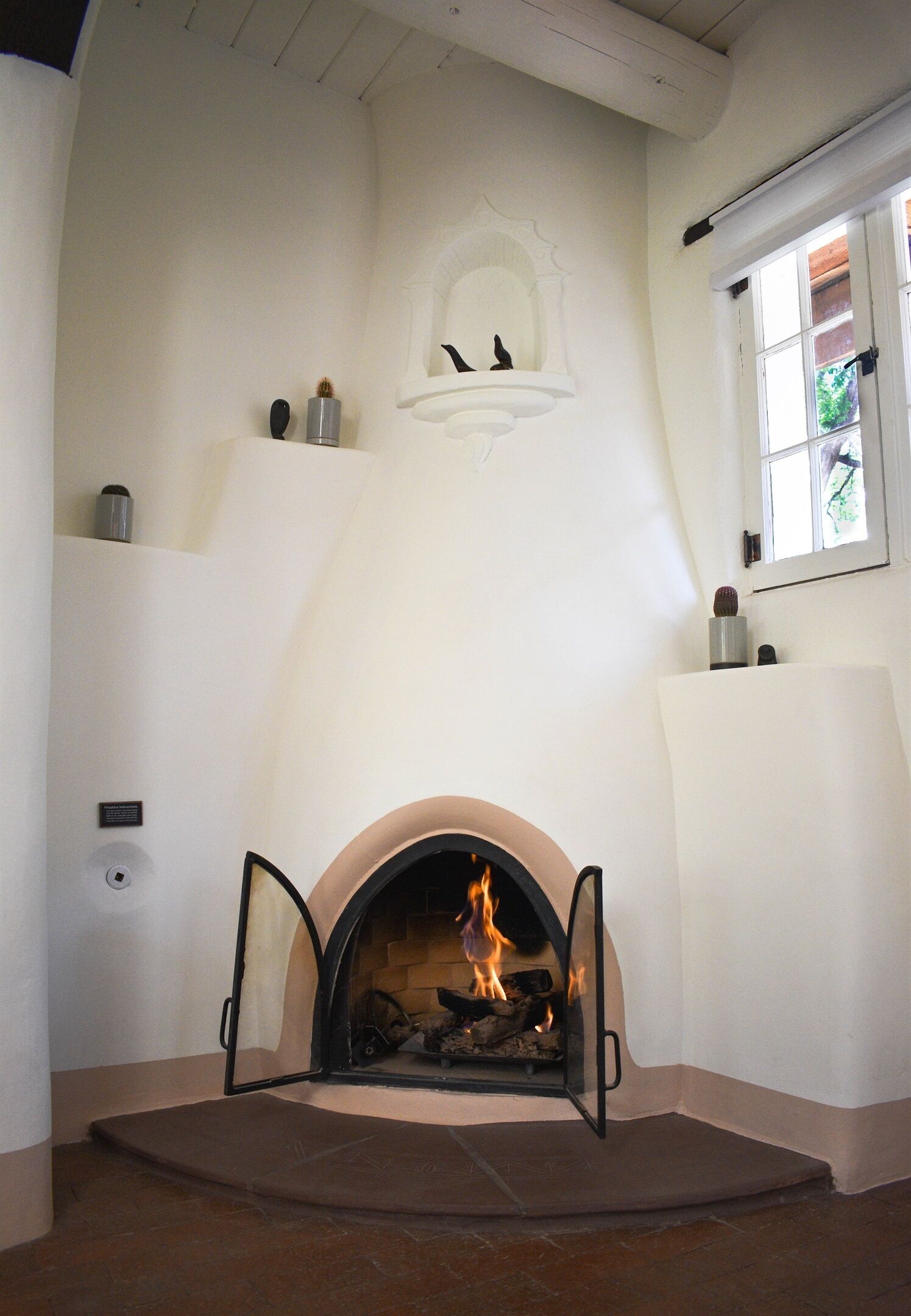 A fireplace with an open door, a wall-mounted shelf with decorative items, and a window with a view of greenery.