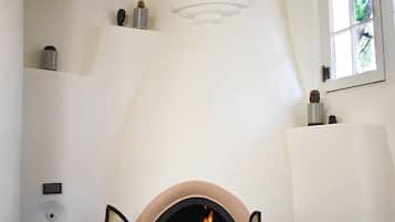 A fireplace with an open door, a wall-mounted shelf with decorative items, and a window with a view of greenery.