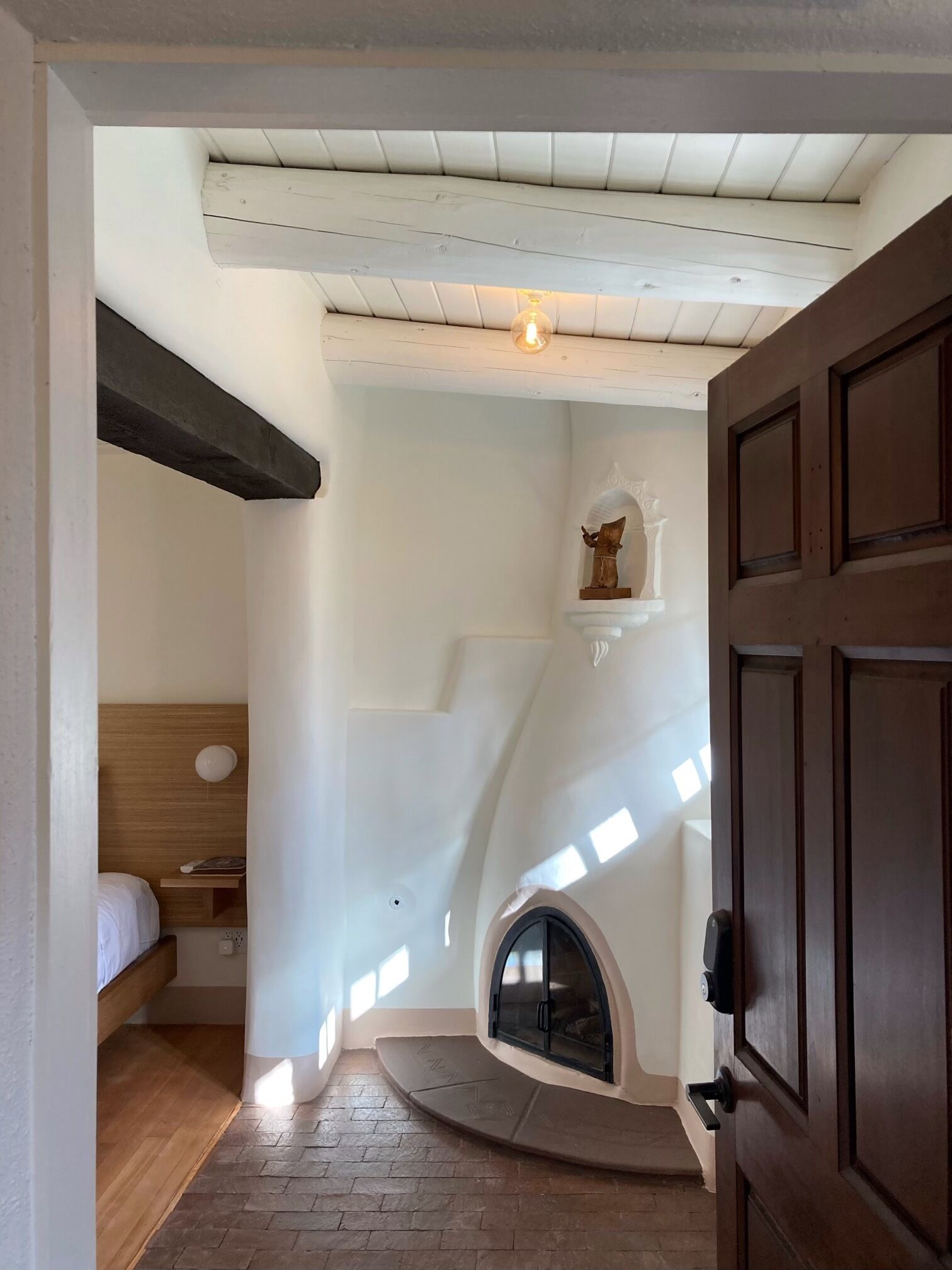 A narrow hallway with a wooden door, a small alcove with a bed, and a fireplace.