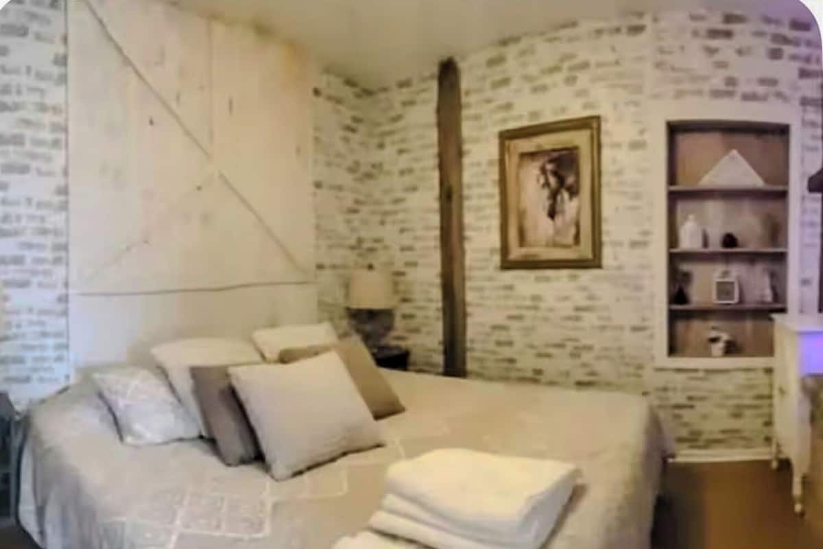 Un dormitorio con una cama grande, una mesita de noche, una pared con un cuadro enmarcado y una estantería con objetos.