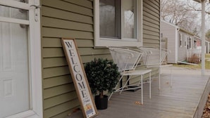 A wooden welcome sign on a porch with white metal chairs and a potted plant.