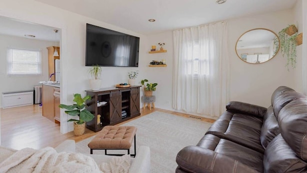 A modern living room with a large flat-screen TV, a leather sofa, and a small ottoman. There are potted plants and a round mirror on the walls.