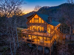 Una baita di legno con un'ampia veranda e numerose finestre, immersa tra alberi con montagne sullo sfondo.