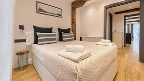 A bedroom with a large bed, a wooden beam ceiling, a framed picture on the wall, and a small stool. - Modern and Luxurious in Historic Old Town (Bilbao)