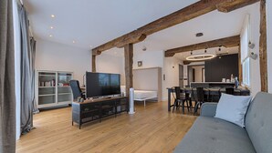 A modern living room with a wooden floor, a large flat-screen TV, a sofa, a dining area with chairs, and a kitchen in the background. - Modern and Luxurious in Historic Old Town (Bilbao)