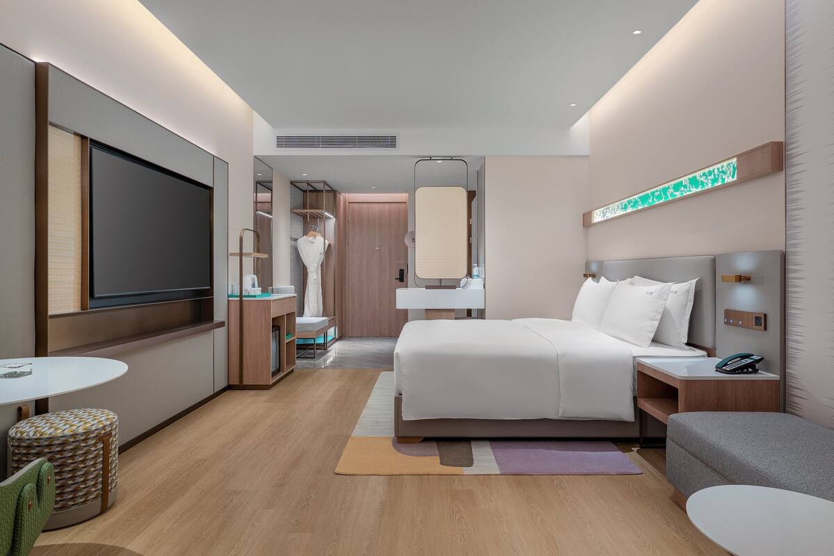 A modern hotel room with a large bed, bedside tables, a TV, a seating area, and a bathroom visible through an open door.