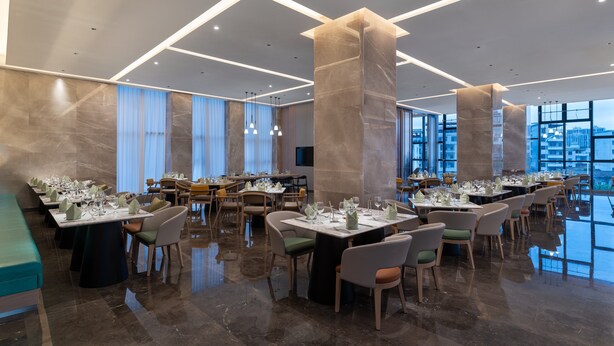 A modern restaurant with a high ceiling, marble flooring, and large windows offering city views. Tables are set with white tablecloths and green napkins.