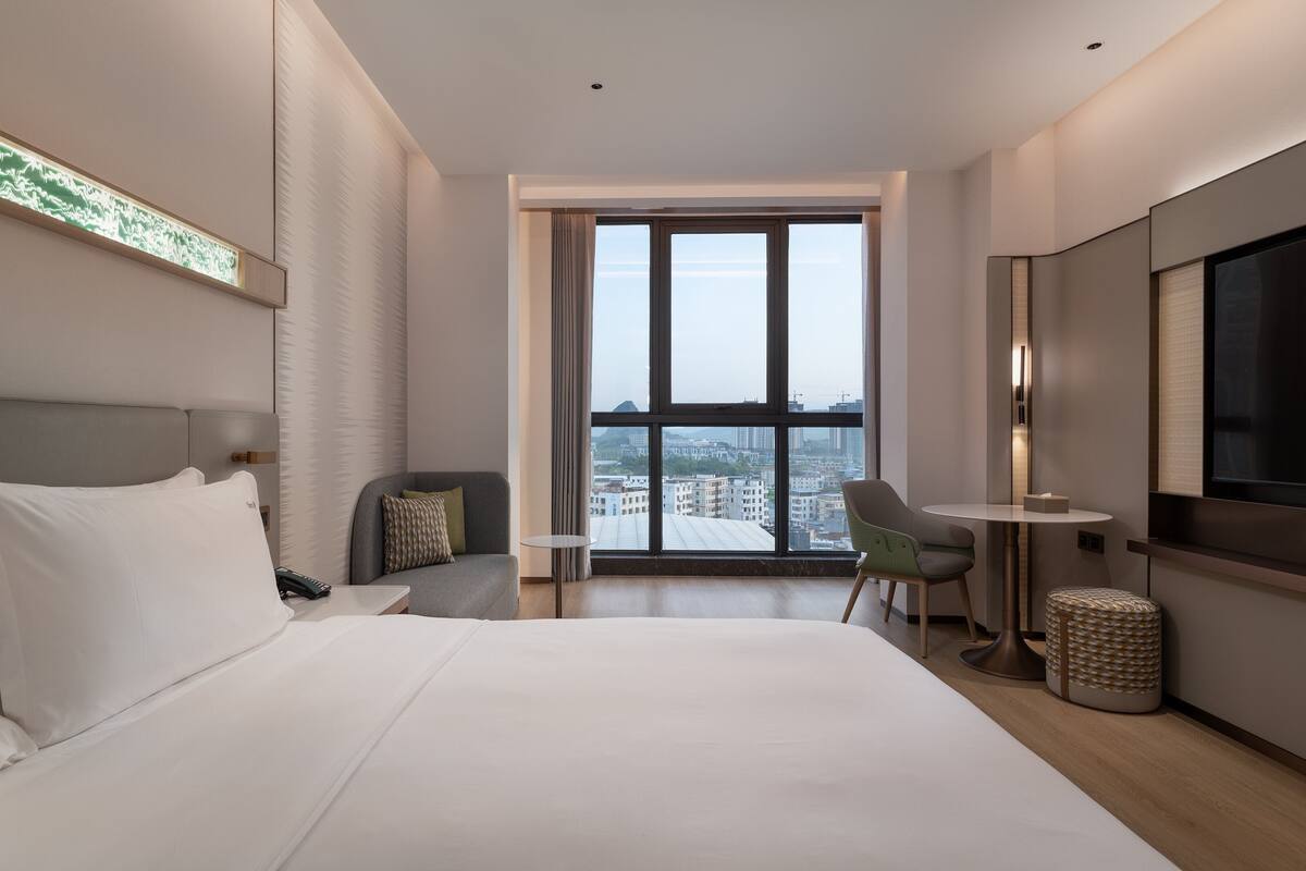 A modern hotel room with a large bed, a TV, a desk, and a view of the city.