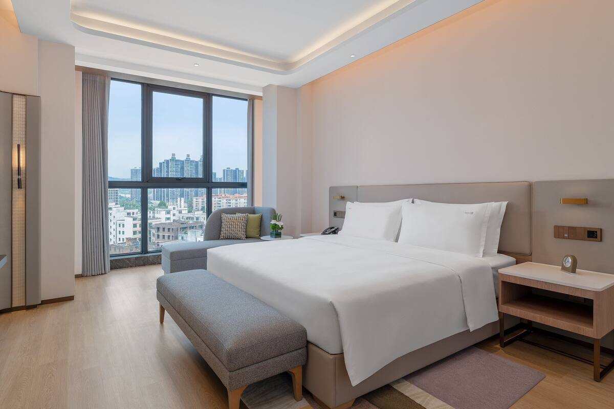 A modern hotel room with a large bed, bedside tables, a bench, a TV, and a view of the city through large windows.