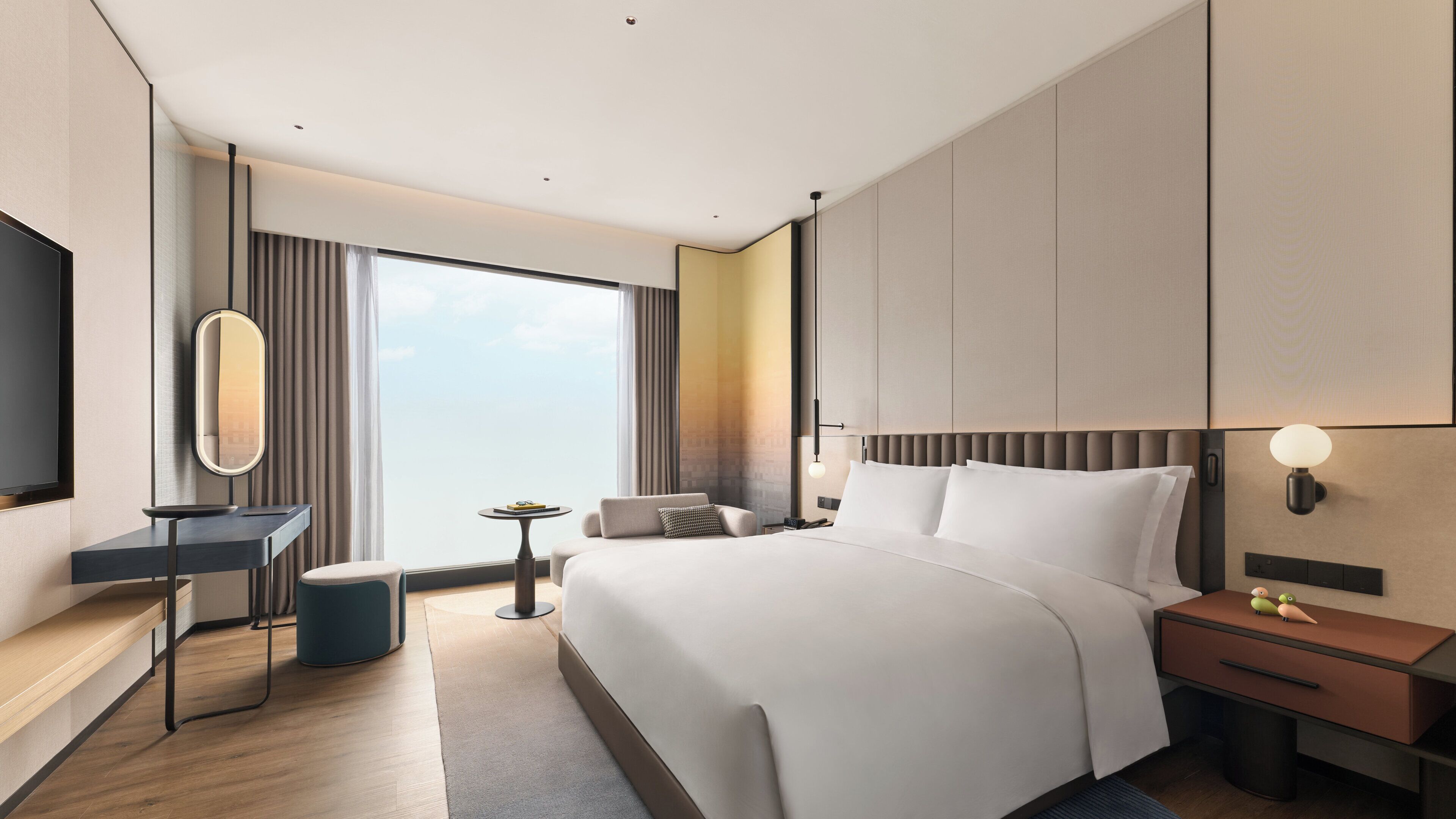 A modern hotel room with a large bed, bedside tables, a desk, a chair, a small table, and a large window with curtains.