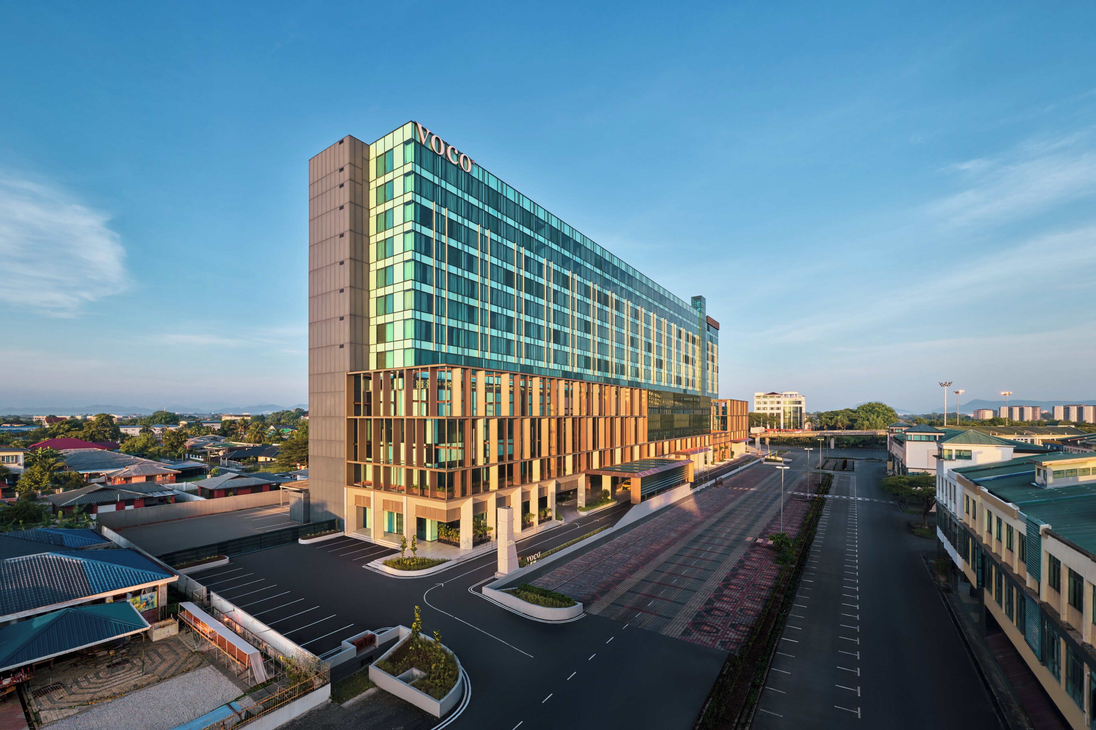 A modern hotel with a prominent 'Voco' sign, featuring a glass facade, a spacious parking area, and a view of the surrounding urban landscape.
