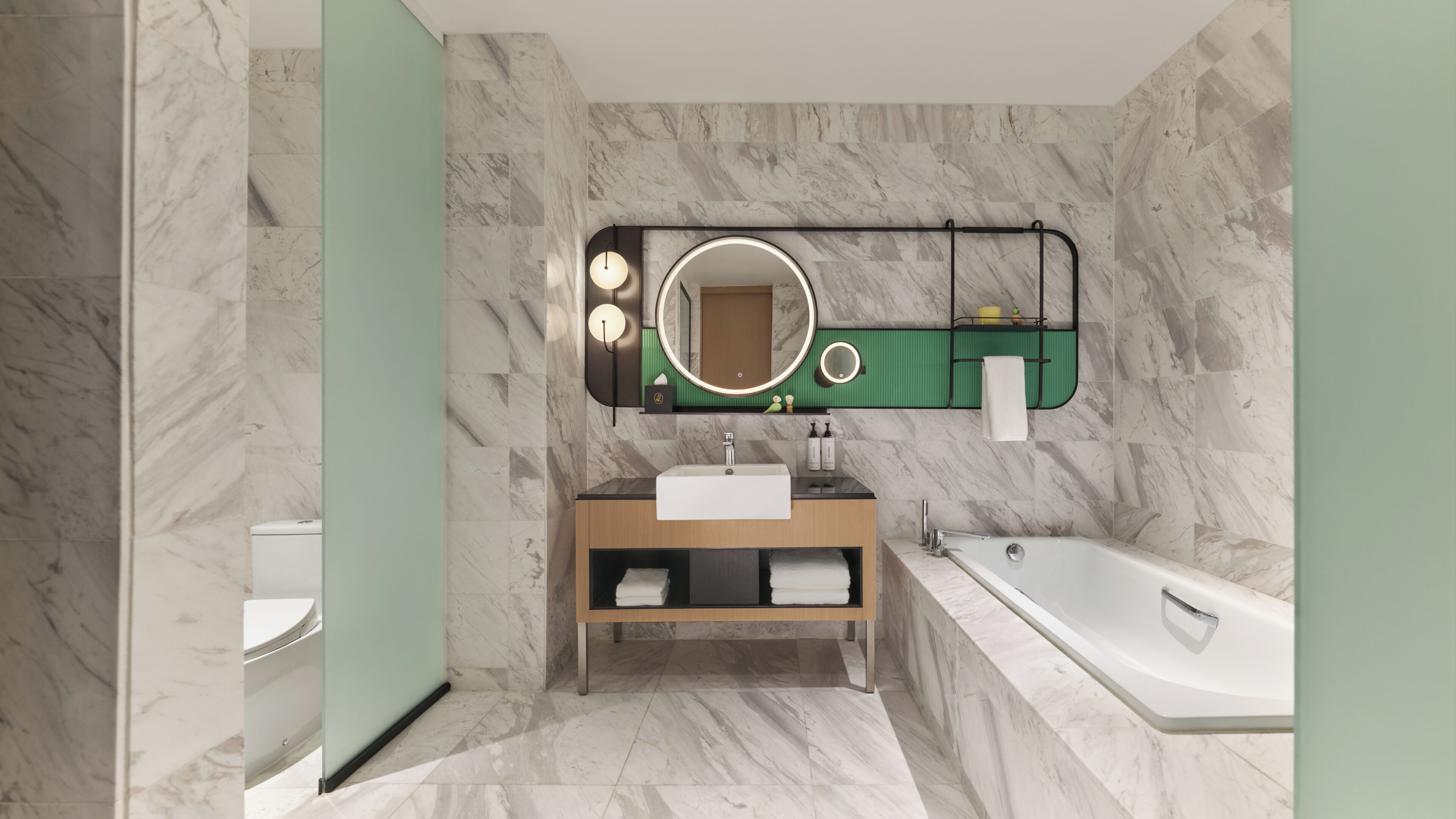 A modern bathroom with a marble floor, a freestanding bathtub, a vanity with a mirror, and a green shelf above the mirror.
