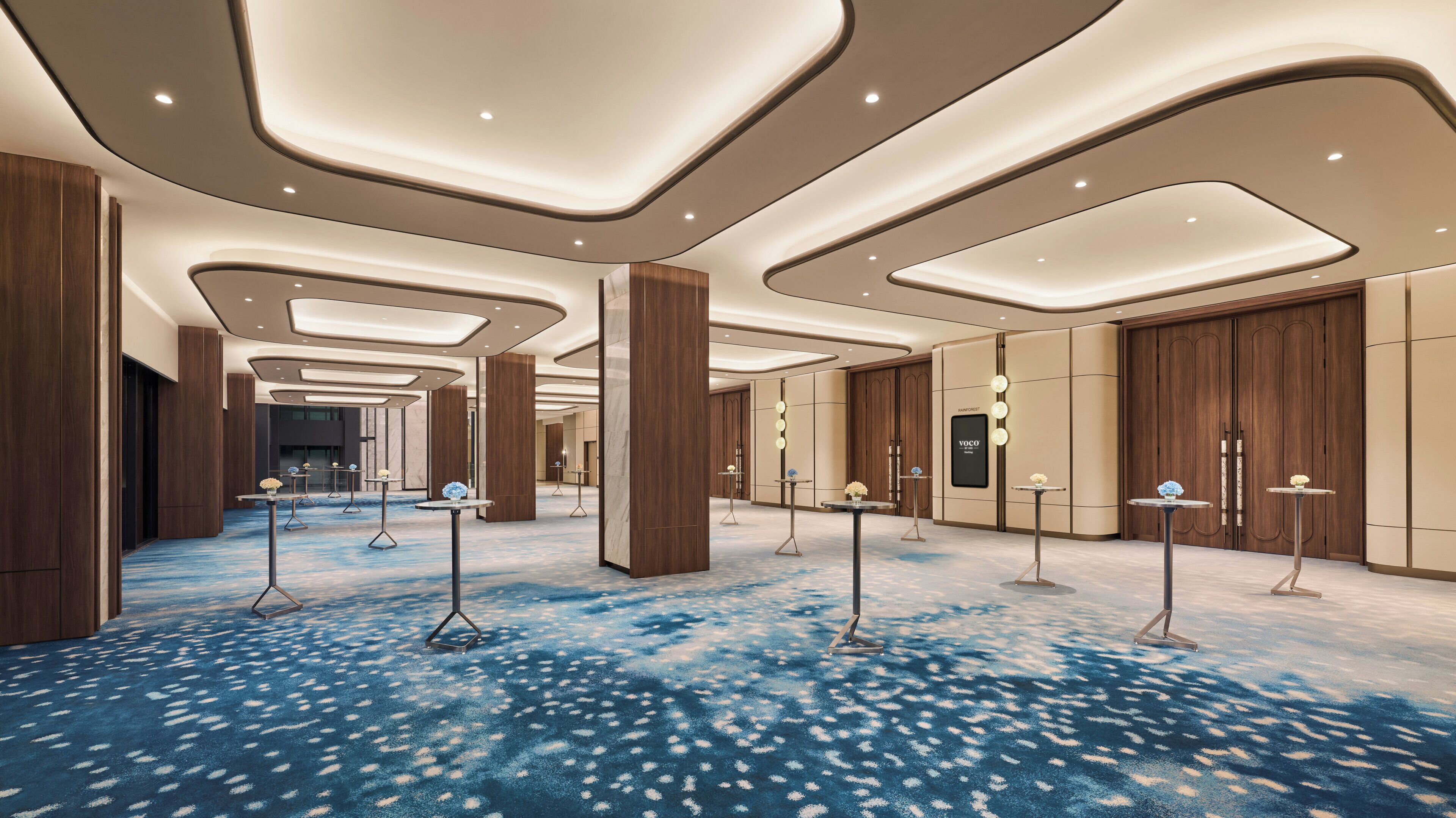Un amplio hall de hotel con diseño moderno, que presenta una alfombra azul estampada, paneles de madera y luminarias contemporáneas.