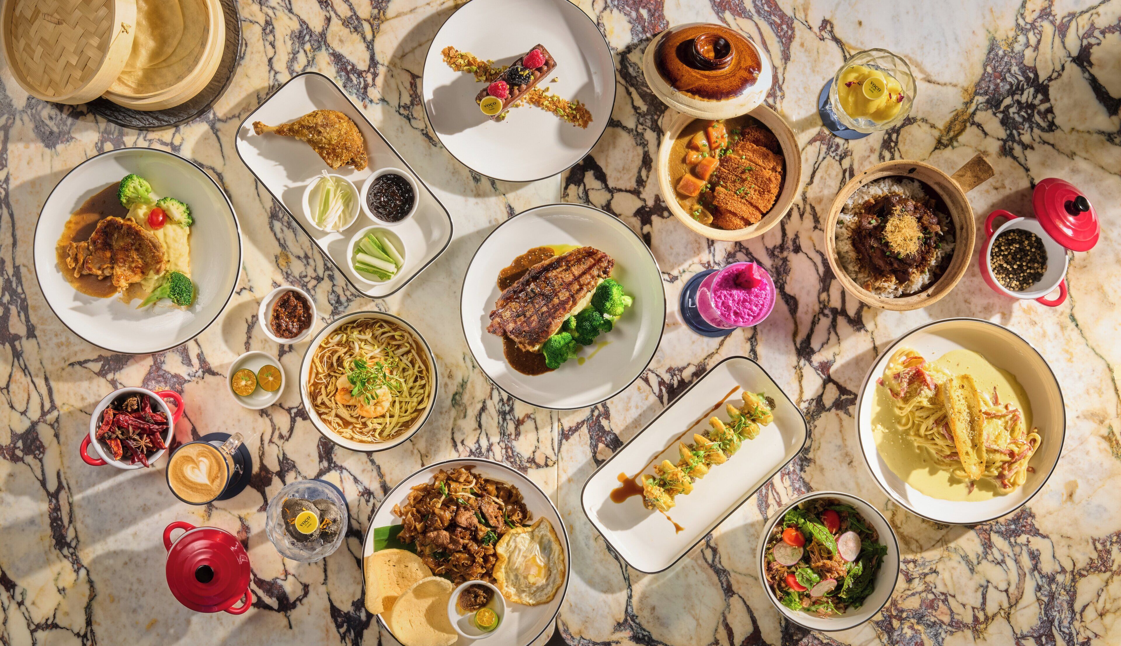 A variety of dishes on a marble table, including a bowl of noodles with shrimp, a plate of fried chicken, and several desserts.