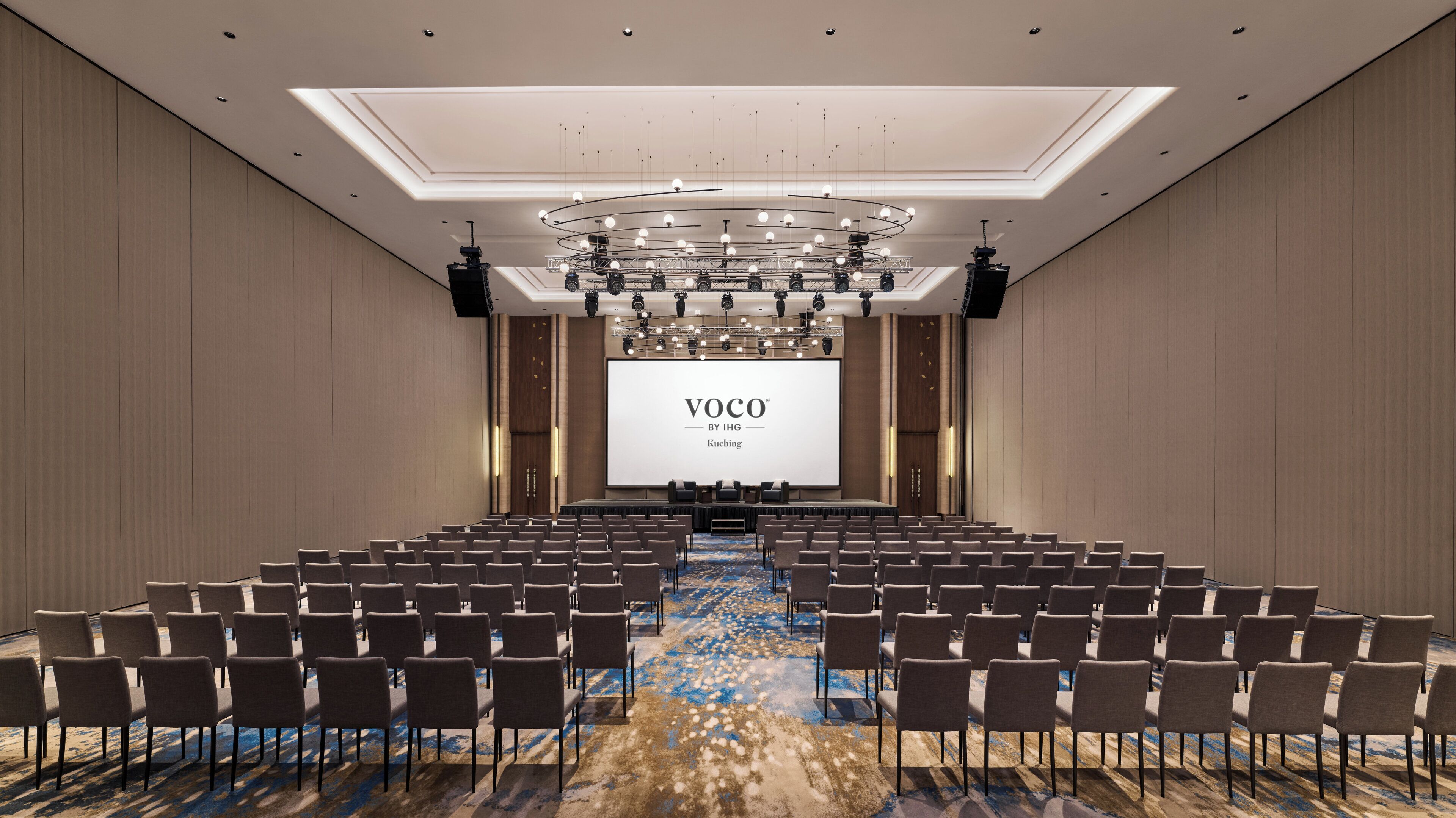 A conference room with rows of chairs, a large screen displaying 'VOCO BY IHG Kuching', and a modern ceiling design.