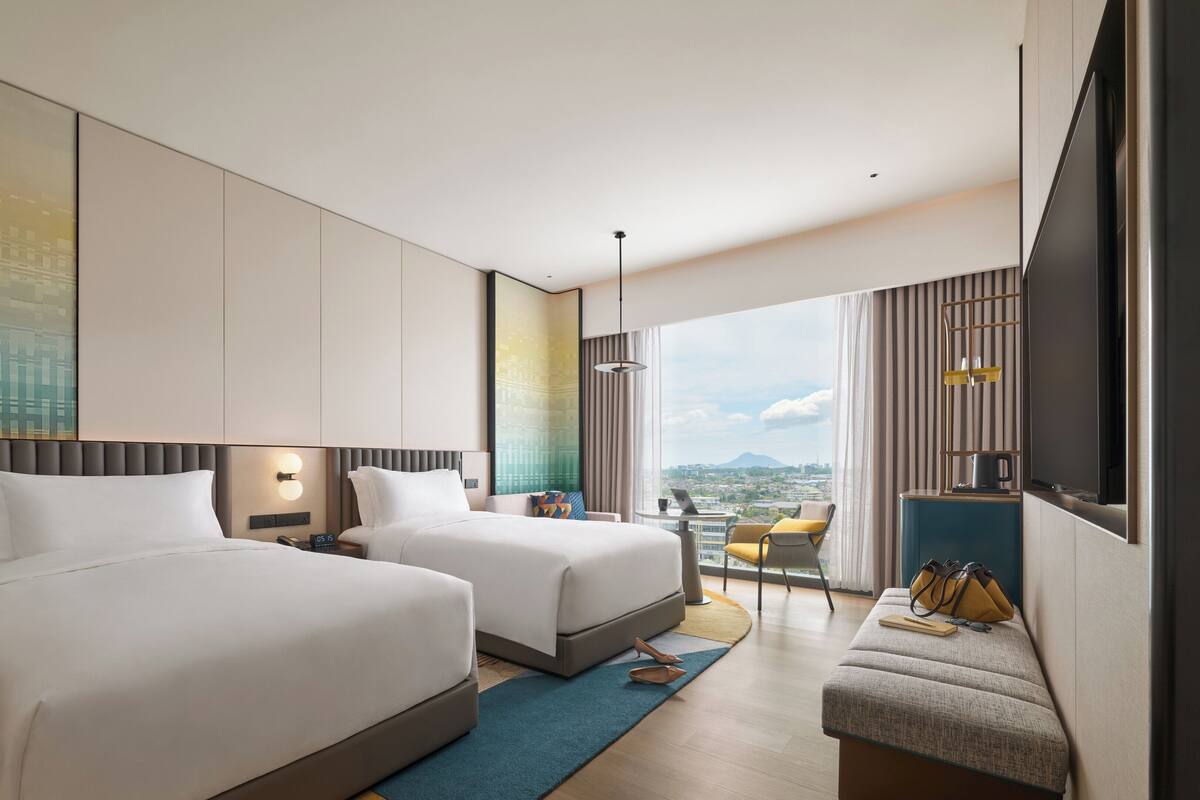 A modern hotel room with two beds, a desk, a chair, and a large window with a city view.