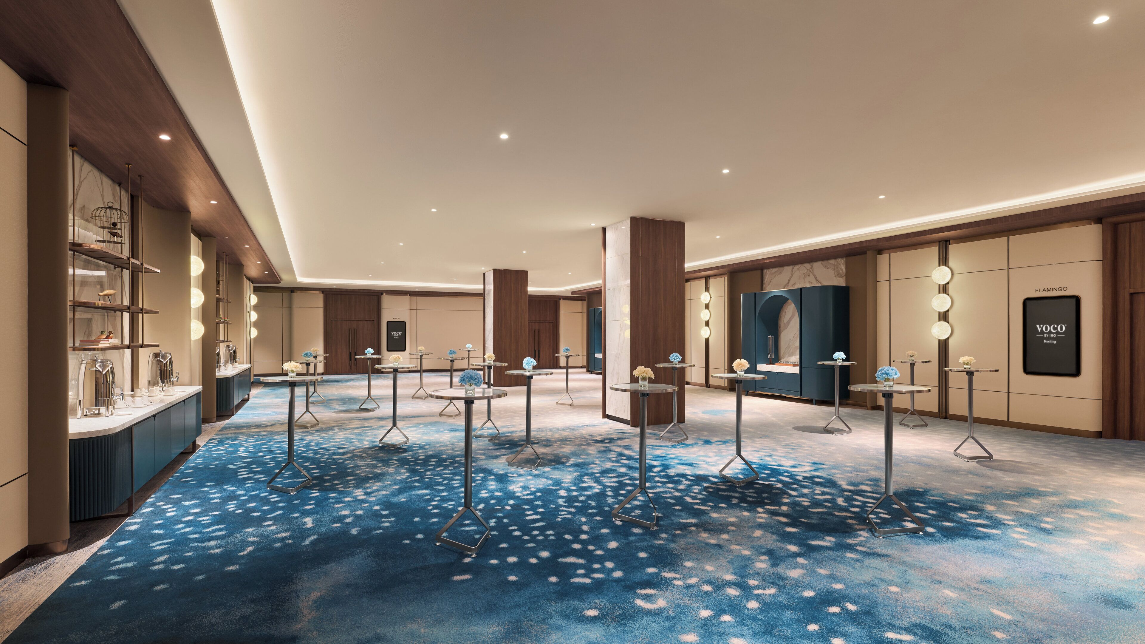 Eine großzügige Hotelhalle mit blauem, gemustertem Teppich, mehreren runden Tischen mit floralen Arrangements und einer großen, dunkel gefärbten Vitrine, an deren Wand das Wort 'FLAMINGO' steht.