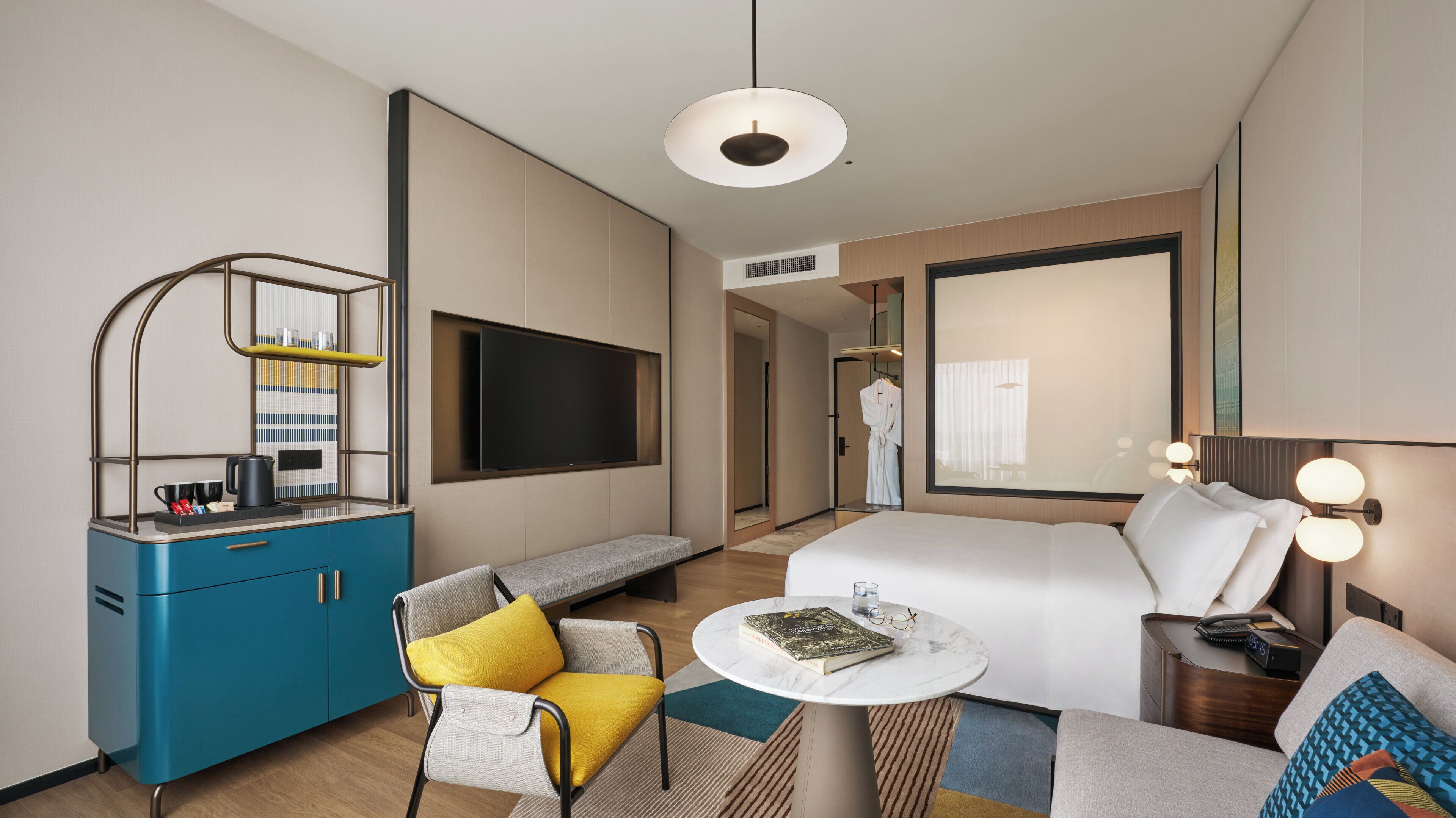 Une chambre d’hôtel moderne comprenant un lit, un coin salon avec un canapé et un fauteuil, une petite table ronde, un téléviseur fixé au mur et une armoire.