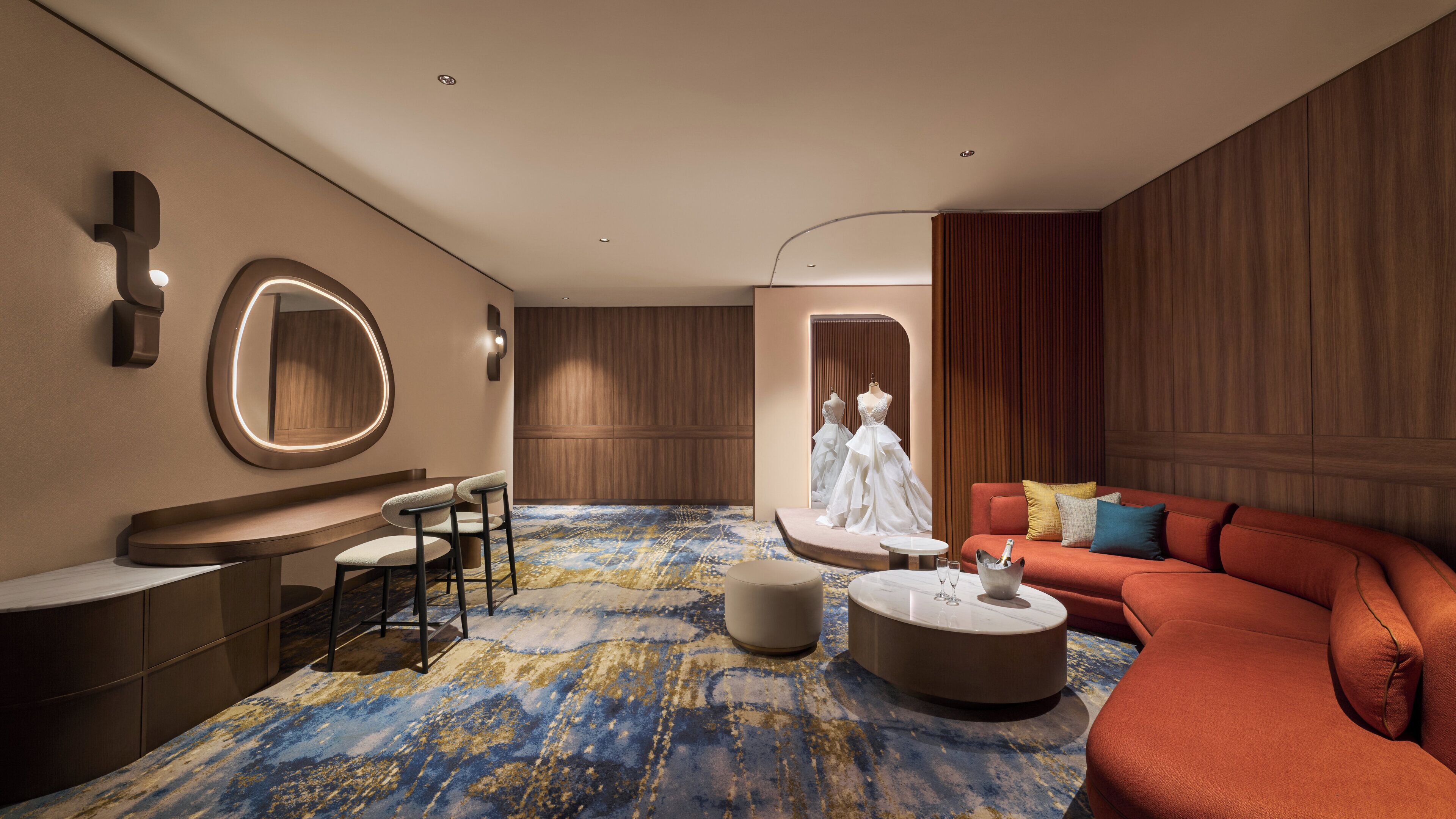 A modern hotel room with a seating area, a large mirror, a dress display, and a red sofa.