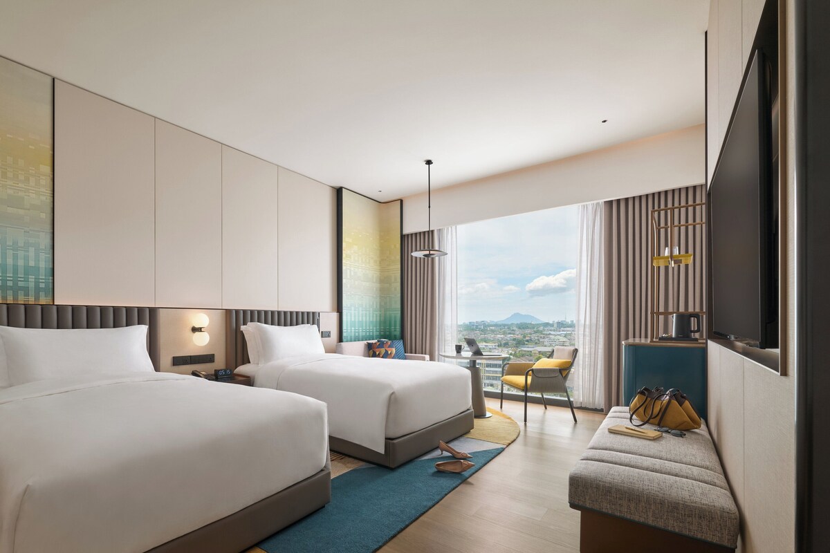 Ein modernes Hotelzimmer mit zwei Betten, einem Schreibtisch, einem Sessel und einem großen Fenster mit Blick auf die Stadt.