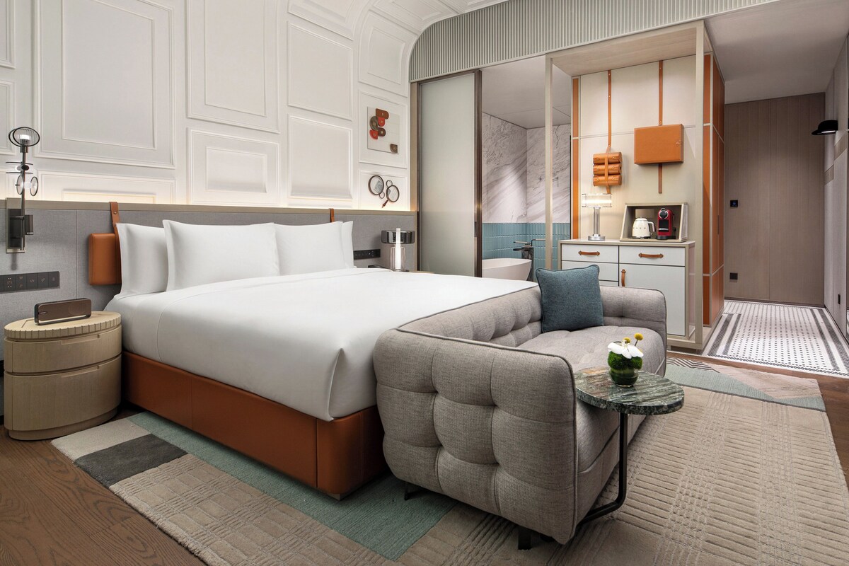 Habitación de hotel moderna con una cama grande, un sofá, una mesita con un florero, y un baño visible a través de una puerta abierta.