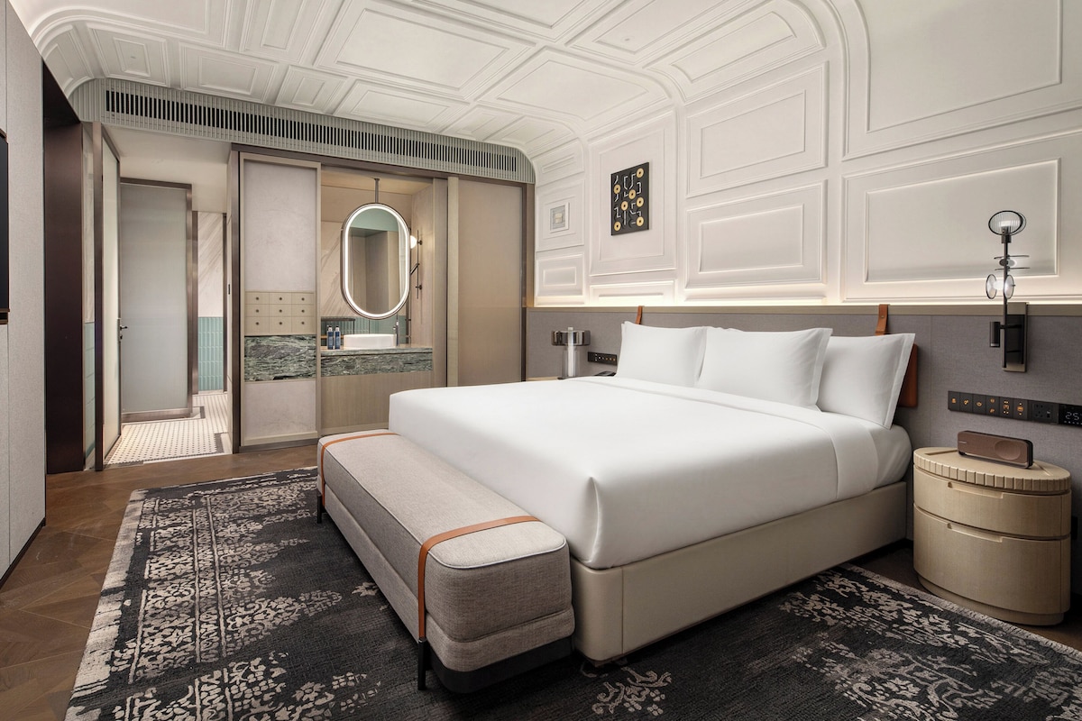 Una habitación de hotel moderna con una cama grande, una mesita de noche, un reloj de pared y una pequeña mesita con una lámpara.